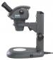 Preview: The image shows a dark-colored microscope with a thick, round head and a magnifying glass. It stands on a sturdy base with an illuminated area and a switch.