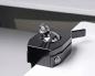 Preview: The image shows a black clamping mechanism with a silver, round head. The mechanism is positioned on the edge of a table and has a lever for tightening.