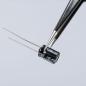 Preview: The image shows a black electrolytic capacitor with two long, thin wires. It is being held with tweezers and has a label on the side. The background is light.