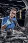 Preview: A young man is wearing safety goggles and gloves while working on a vehicle. He is checking cable connections in a busy workshop environment.