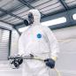 Preview: A person is wearing a white jumpsuit and a breathing mask. They are holding a hose with a spray device, ready to work in a bright, modern room.