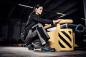 Preview: A sportily dressed woman is kneeling at a yellow device in an industrial hall. She is wearing gray work clothes and looking forward intently. The environment is modern and functional.