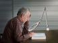 Preview: An elderly man with gray hair and a plaid shirt sits at a table, looking intently into a book. A magnifying glass and a lamp assist him in reading.