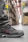 Preview: Das Bild zeigt einen Fuß, der einen schwarzen Sicherheits-Schuh trägt, mit roten Akzenten. Im Hintergrund sind Regale mit Kisten und eine breite Lagerhalle zu sehen.