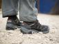 Preview: Das Bild zeigt die Füße einer Person in robusten, schwarzen Arbeitsschuhen auf einem steinigen Untergrund. Die Hosenbeine sind weit und grau, und es gibt farbige Akzente an den Schuhen.