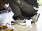 Preview: Eine Person sitzt auf dem Boden, trägt schwarze Arbeitshosen und graue, sportliche Schuhe. Werkzeuge liegen verstreut auf dem Boden, die Umgebung wirkt wie eine Werkstatt.