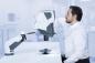 Preview: A man is sitting at a table, intently looking at a medical device with a movable arm. The device has a white surface and is located in a bright room.