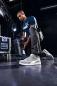 Preview: A man in a dark blue T-shirt with the Puma logo bends down and picks up a black lamp. He is wearing gray sneakers and dark, loose pants. The background is modern and industrial.