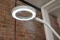 Preview: The image shows a white reading lamp with a circular LED ring and a magnifying lens in the center, mounted on a flexible arm. In the background, there is a brick wall.