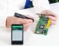Preview: In the image, one hand is holding a circuit board while the other hand is using a measuring device. Next to the hand, there is a smartphone displaying values. The background is bright and neutral.