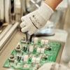 Preview: A hand in a white glove holds a pair of pliers and works on a green circuit board with various components on it. In the background, an industrial environment is visible.