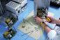 Preview: In the picture, you can see a work desk with technical equipment. A person is holding a screwdriver and working on an open case to repair electronic components. Around them, screws and tools are scattered.