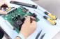 Preview: In the image, you can see a manual assembly or repair of a green circuit board with many electronic components. Next to it, there are screwdrivers and pliers.