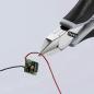 Preview: A pair of pliers holds two wires, a red one and a black one, which are attached to a small green circuit. The background is bright and neutral.