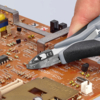 Preview: Auf dem Bild sieht man eine Hand, die eine Zange hält. Damit wird eine elektronische Platine bearbeitet, die viele kleine Bauteile und Leiterbahnen auf einer braunen Oberfläche hat.