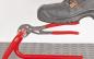 Preview: A black work shoe is standing on a red pliers, which is lying on a gray rubber floor. The pliers are open and have red handles. The wall in the background is white.