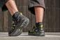 Preview: Auf dem Bild sind die Füße einer Person in sportlichen, schwarzen Schuhen mit gelben Akzenten zu sehen. Die Hose ist kurz und der Hintergrund besteht aus einer Holzstruktur.
