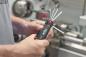 Preview: The image shows a person holding a compact toolset in their hand. The handle is black with red accents. Several metal rods protrude from the top end, resembling hex keys. In the background, machines and a workshop can be seen.