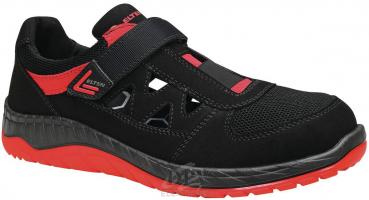 Der Schuh ist ein sportlicher Halbschuh in Schwarz mit roten Akzenten. Er hat einen breiten Klettverschluss und offene Stellen an der Seite für Belüftung. Die Sohle ist rutschfest und leicht.