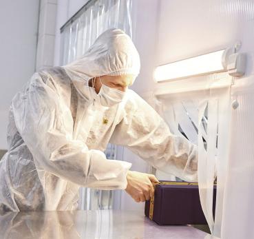 Eine Person in einem weißen Schutzhanzug und Maske arbeitet an einem Tisch. Sie öffnet eine lila Kiste. Der Raum wirkt steril, mit hellem Licht und durchsichtigen Vorhängen.