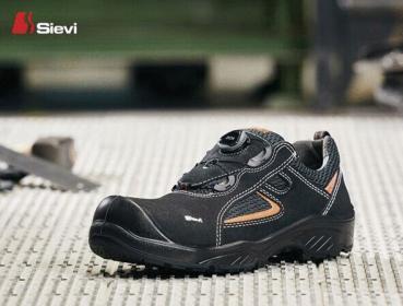 Das Bild zeigt einen schwarzen Arbeitsschuh mit runder Zehenkappe. Er hat orangefarbene Akzente und eine rutschfeste Sohle. Der Schuh ist auf einem grauen, strukturieren Untergrund platziert.
