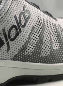 Das Bild zeigt einen sportlichen Schuh in Grau und Weiß mit einem modernen, strukturierten Obermaterial. Der Markenname ist seitlich in Schwarz aufgedruckt. Die Sohle ist dunkel und rutschfest.