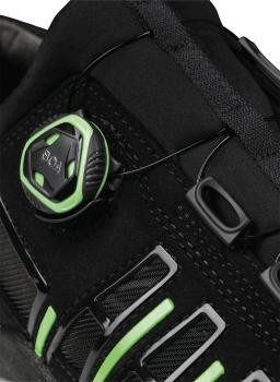 Das Bild zeigt einen Teil eines sportlichen Schuhs in Schwarz mit grünen Akzenten. Ein runder Verschlussmechanismus ist prominent, um die Schnürung anzupassen. Es gibt auch Seitenschlitze.