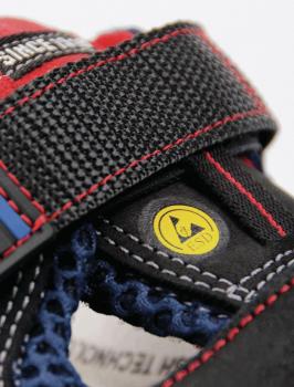 The image displays a detailed section of a shoe. It is black with red and blue accents. A wide strap made of textured material holds the shoe together, and a yellow logo is visible.