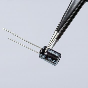 The image shows a black electrolytic capacitor with two long, thin wires. It is being held with tweezers and has a label on the side. The background is light.