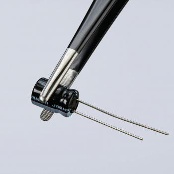 The image shows a pair of pliers holding a small, round, black component with two protruding wires. The component has a smooth surface and bears an inscription.