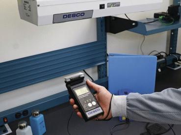 The image shows a handheld device from DESCO being held in hand. In the background, shelves with various equipment and a blue binder are visible.