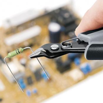 Auf dem Bild sieht man eine Hand, die eine Zange hält, die einen Widerstand greift. Im Hintergrund ist eine Leiterplatte mit verschiedenen elektronischen Bauteilen unscharf zu erkennen.