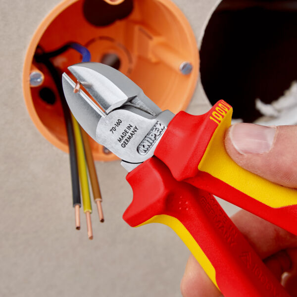 The image shows a hand holding a red and yellow pliers. The pliers are used for cutting wires near an electrical junction box with multiple cables.