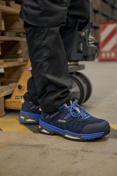 Das Bild zeigt einen stehenden Mann in einer dunklen Hose, der Sicherheitsschuhe in Blau und Schwarz trägt. Der Hintergrund ist ein Lager mit Holzpaletten.