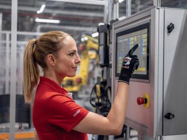 A woman in a red shirt is operating a touchscreen on a machine. She is wearing a black glove and looking intently at the screen. Robots are visible in the background.