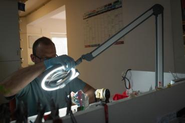 Der Bildausschnitt zeigt eine Person mit Handschuhen, die konzentriert unter einer hellen, runden Arbeitslampe an einem Tisch arbeitet. Im Hintergrund ist eine Wand mit einem Kalender sichtbar.