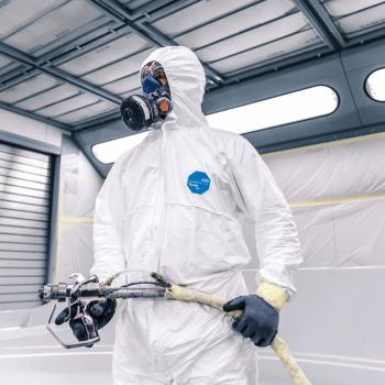 A person is wearing a white jumpsuit and a breathing mask. They are holding a hose with a spray device, ready to work in a bright, modern room.