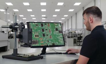 Ein Mann sitzt an einem Tisch und betrachtet auf einem Monitor eine grüne Leiterplatte mit Bauteilen. Der Raum ist hell und modern, mit weiteren Arbeitsplätzen im Hintergrund.