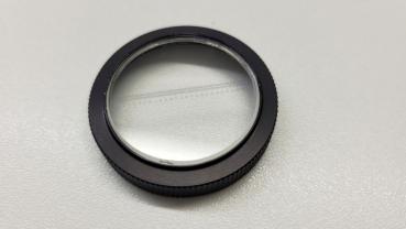 The image shows a round, flat lens frame made of black metal. In the center, there is a clear, transparent lens. The edge has a ridged structure for better grip.