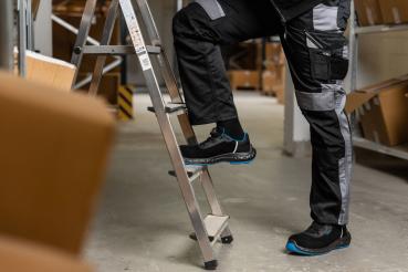 Auf dem Bild sieht man einen Mann, der auf einer Leiter steht. Er trägt schwarze Arbeitskleidung mit grauen und blauen Akzenten. Im Hintergrund sind Kartons in einem Lager zu sehen.