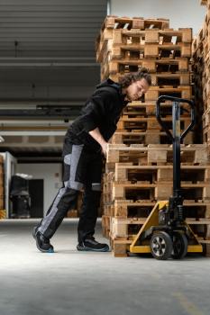 Ein Mann beugt sich über einen Holzstapel aus Paletten. Er trägt eine schwarze Jacke und graue Arbeitshose. Neben ihm steht ein Handwagen mit einer großen Rolle. Helle Halle im Hintergrund.