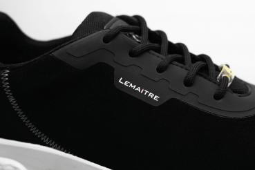 Das Bild zeigt einen schwarzen Sportschuh mit einem glatten Obermaterial. Die Schnürsenkel und eine weiße Gummisohle sind gut sichtbar. Ein kleines, silbernes Logo ist seitlich angebracht.