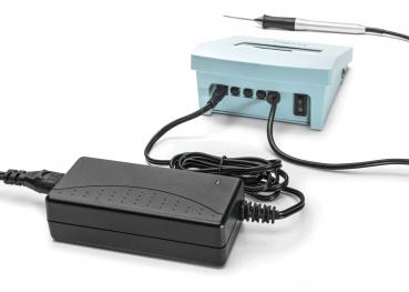 The image shows a device with a light blue box that has cable connections. Next to it is a black power supply with a long cable. A soldering iron is also lying next to it.