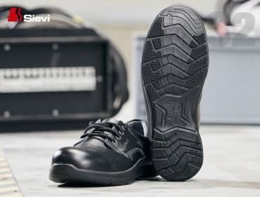 Das Bild zeigt einen schwarzen Sicherheitsschuh von Sievi. Der Schuh hat eine schlichte Schnürung und eine strukturierte, rutschfeste Sohle mit deutlichem Profil. Im Hintergrund sind verschwommene Werkzeuge.
