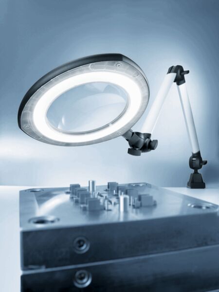 The image shows a magnifying lamp with ring lighting mounted on a table. Below it is a metallic component with various small tools or structures.