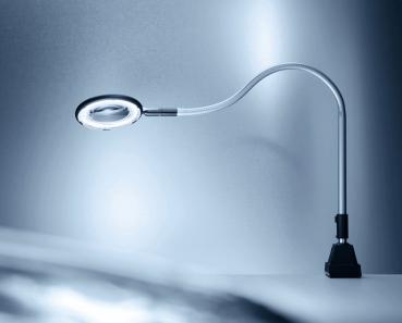 Die Abbildung zeigt eine flexible Schreibtischlampe mit einem runden, vergrößernden Licht. Der Hals der Lampe ist biegsam und sie steht auf einem stabilen, schwarzen Fuß. Der Hintergrund ist in sanften Grautönen.