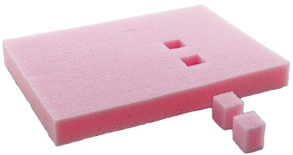 Das Bild zeigt einen rechteckigen, rosa Schaumstoffblock mit zwei kleineren, quadratischen Schaumstoffstücken daneben. Auf der großflächigen Oberfläche sind zwei Vertiefungen.