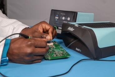 Eine hand arbeitet an einer grünen Leiterplatte, umgeben von technischen Geräten. Ein Gerät in hellem Blau steht daneben, das zur Wartung oder zum Testen genutzt wird.