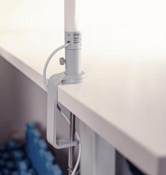 The image displays a white desk lamp attached to the edge of a table. It features a cylindrical shaft and a clamp for stabilization. The background appears bright and neutral.