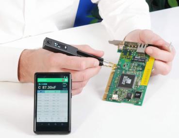 In the image, one hand is holding a circuit board while the other hand is using a measuring device. Next to the hand, there is a smartphone displaying values. The background is bright and neutral.